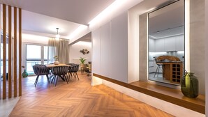 A modern dining area with a wooden herringbone floor, a central dining table with chairs, and a large mirror reflecting the kitchen. - Phaedrus Living: Luxury Penthouse Ploutarchou (Athens)
