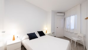 Ein sauberes, weißes Schlafzimmer mit einem Bett, zwei Nachttischen mit Lampen, einem Sessel und einer Klimaanlage.