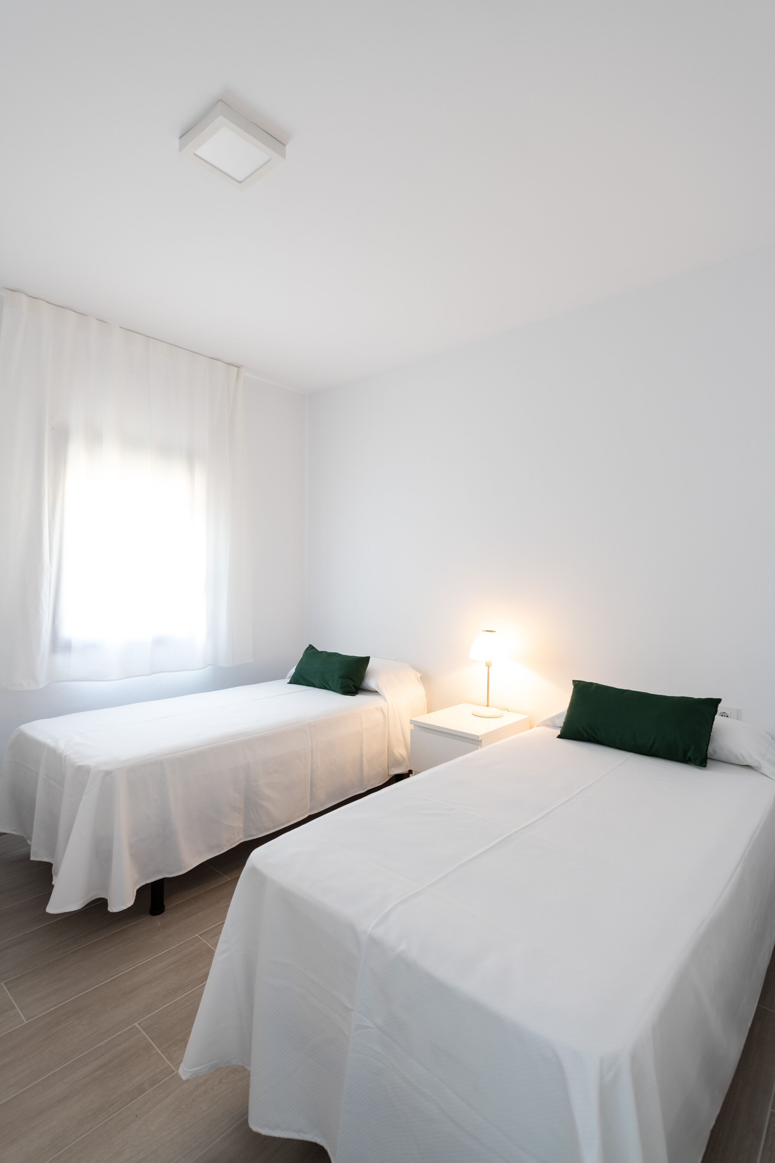 Una habitación limpia y minimalista con dos camas individuales, una mesita de noche con lámpara y una ventana con cortinas translúcidas.
