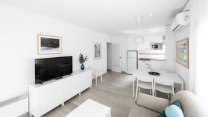 Ein modernes, weißes Wohnzimmer mit Flachbildfernseher, einem Essbereich mit weißem Tisch und Stühlen sowie einer Küchenzeile mit Mikrowelle und Kühlschrank.