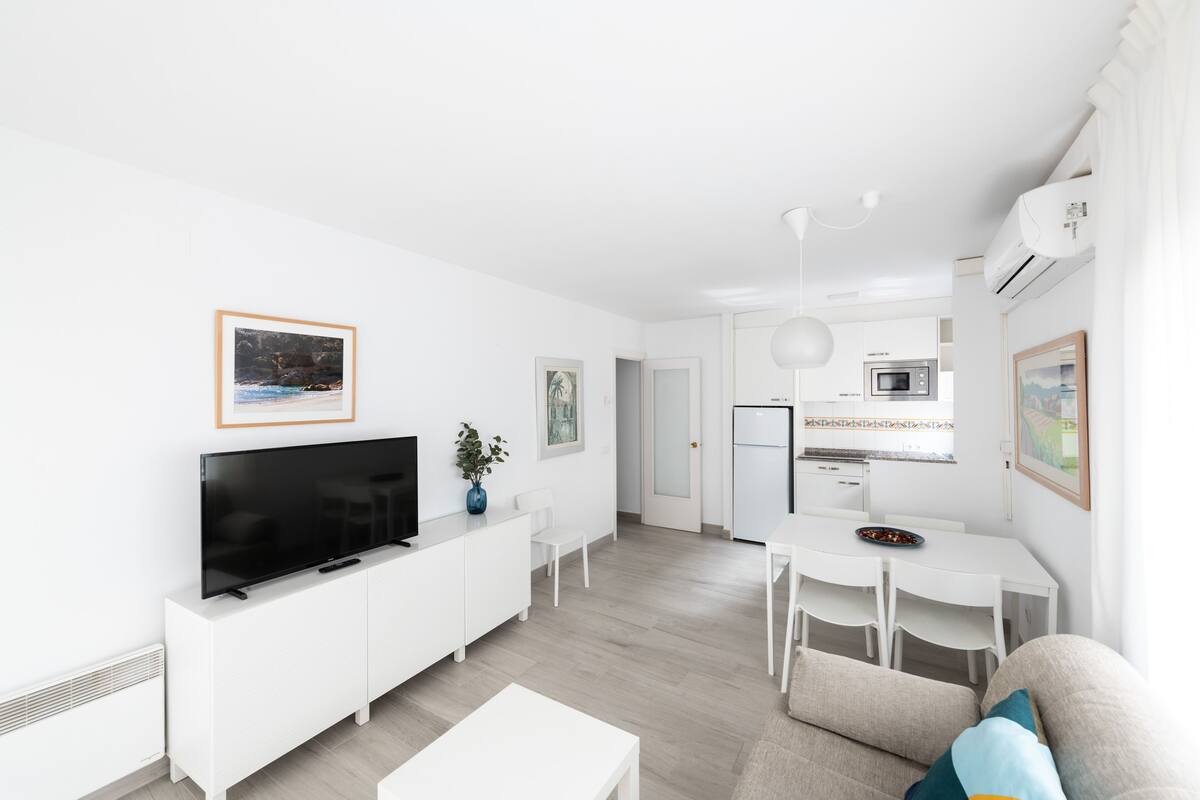 Uma sala de estar moderna, com decoração em branco, que inclui uma TV de tela plana, um espaço de jantar com mesa e cadeiras brancas, e uma cozinha equipada com micro-ondas e geladeira.