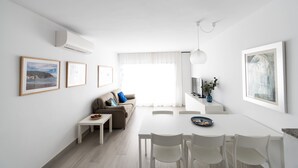 Ein modernes, minimalistisches Wohnzimmer mit einer Couch, einem Esstisch und Stühlen. Ein Fernseher steht auf einem Lowboard und ein Fenster ist mit Vorhängen drapiert.