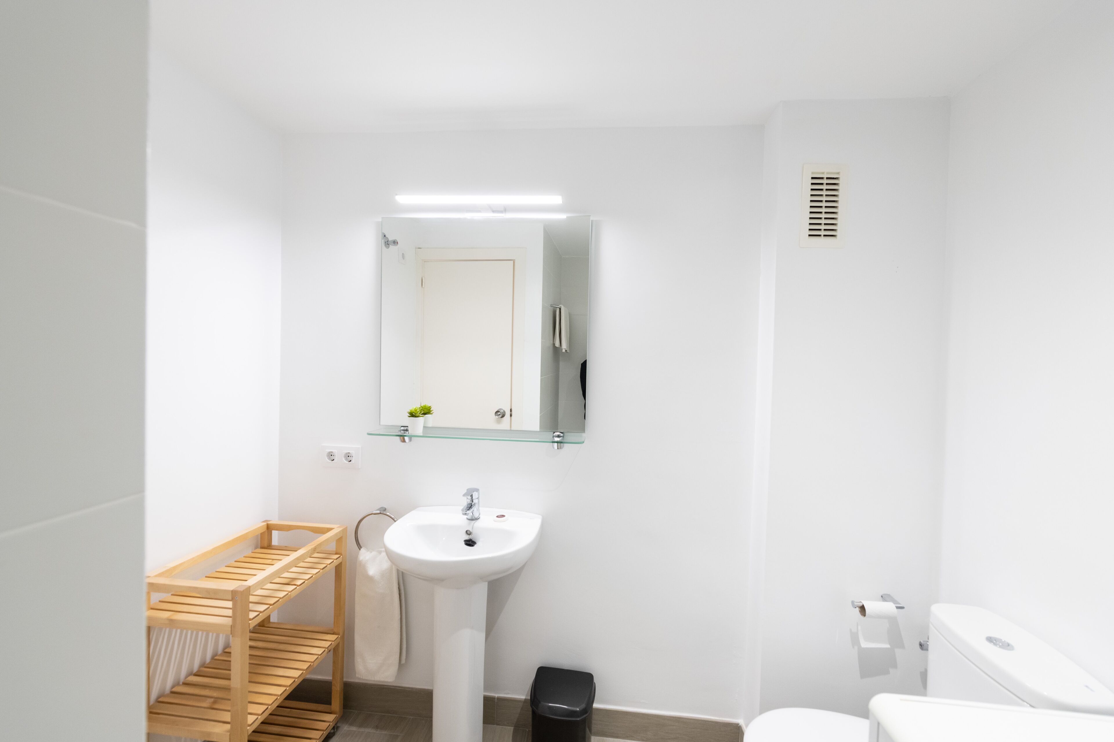 A small, modern bathroom with a white sink, mirror, and toilet. There is a wooden shelf and a black trash bin.