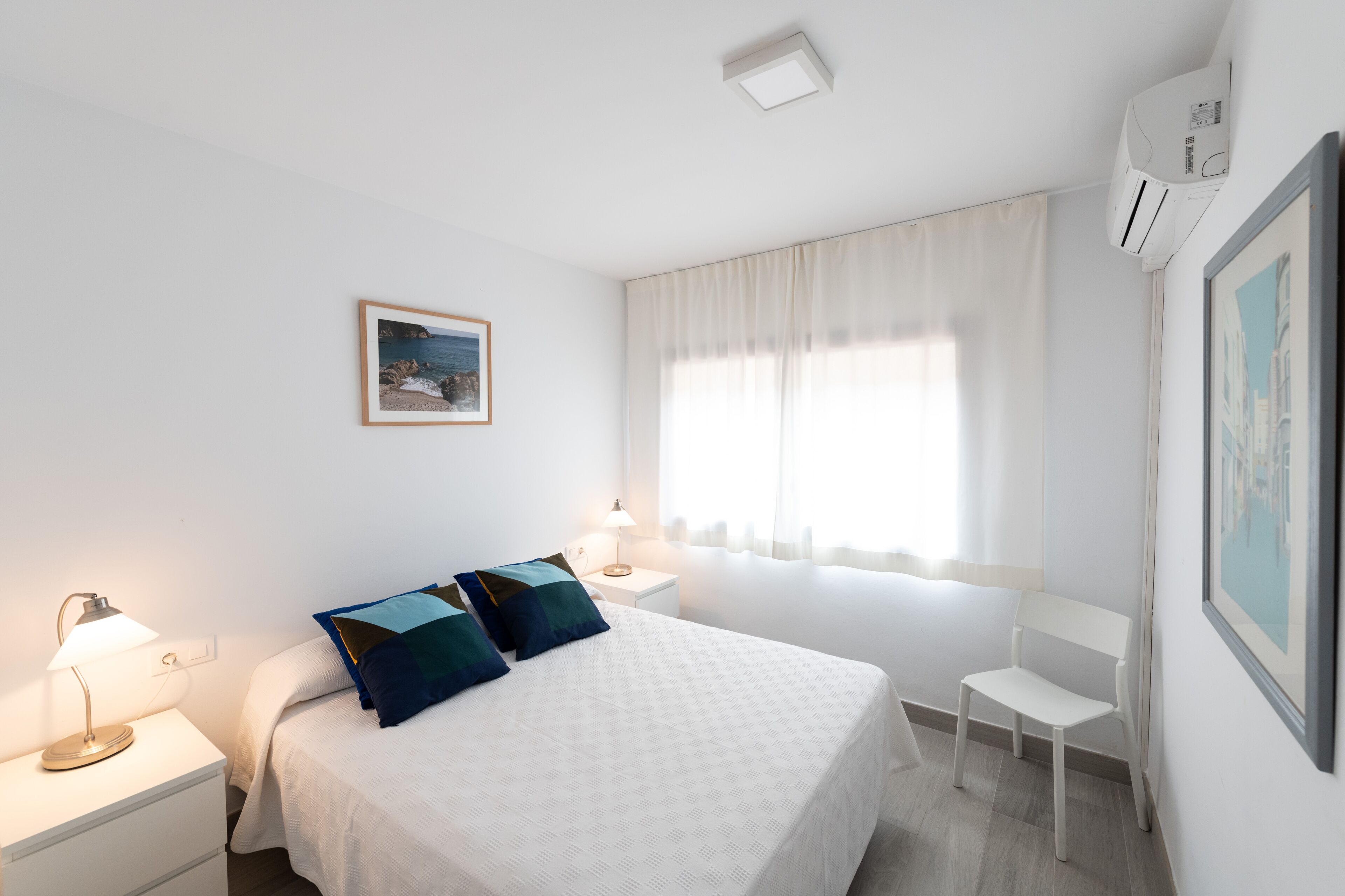 Un dormitorio limpio y minimalista con cama, mesitas de noche, una silla y una ventana con cortinas.