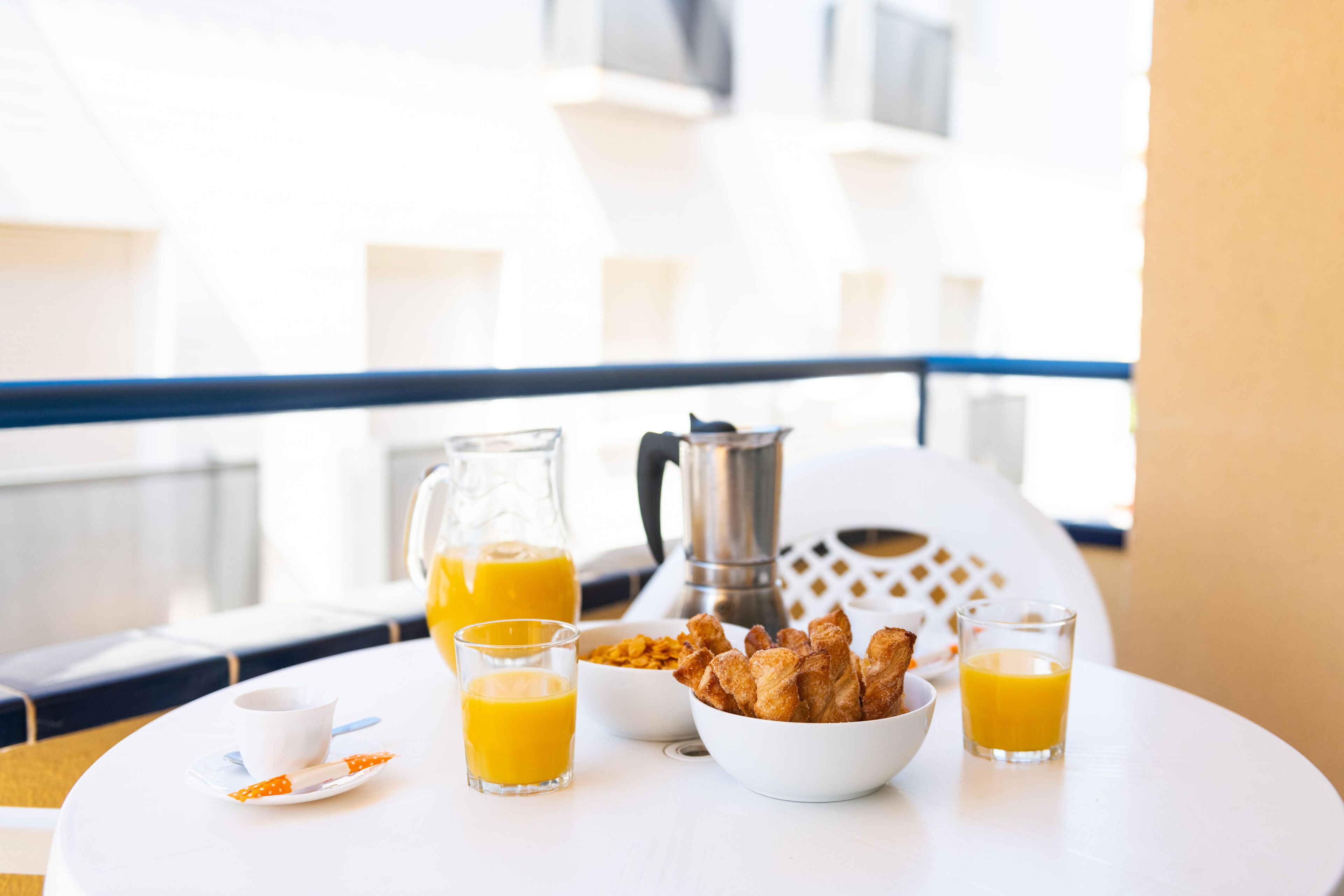 Une table de petit-déjeuner avec du jus d’orange, une cafetière et un bol de viennoiseries, le tout sur un balcon.