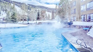 Una piscina con acqua cristallina, circondata da terreno e alberi coperti di neve, con un edificio sullo sfondo.