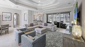 Un salon spacieux avec un canapé d’angle, un pouf et un grand tapis. On y trouve également un coin repas avec une table et des chaises, et une cuisine à l’arrière-plan.