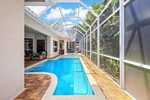 An indoor swimming pool with a glass ceiling, surrounded by a patio area with seating and a view of the outdoors.