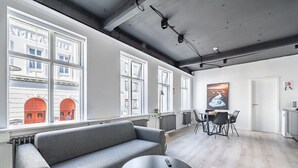 A modern interior with a grey sofa, a round black table, and a dining area with chairs. Large windows provide natural light and a view of buildings with red doors.