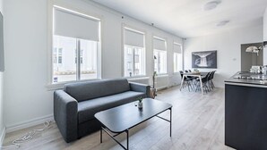 Un appartement moderne avec un canapé gris, une petite table, un coin repas avec des chaises et un coin cuisine équipé d’un évier et d’un plan de travail.
