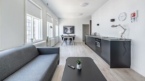 Un appartement moderne avec un canapé gris, une petite table agrémentée d’une plante, un coin cuisine et une table à manger avec des chaises.