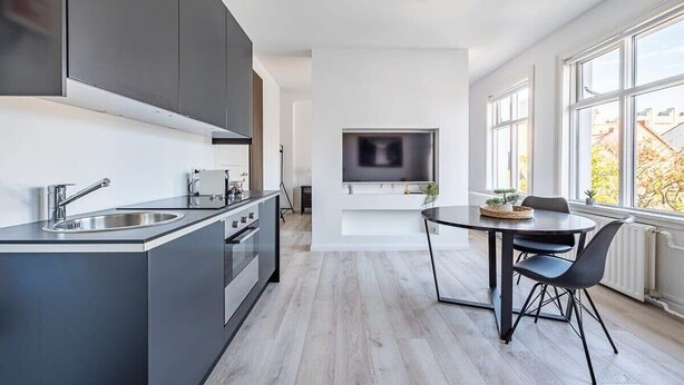 Una cuina moderna amb una illa central, una zona de menjador amb taula i cadires, i un gran finestral que ofereix llum natural.