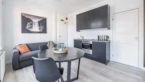 Una cuina moderna amb armaris foscos, una zona de menjador amb taula rodona i cadires, i un sofà gris amb coixins taronja.