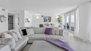 Un salon moderne avec un grand canapé en forme de L, une table basse et un lampadaire. Un tableau est accroché au mur et une plante est posée sur le rebord de la fenêtre.