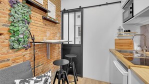 A compact kitchen with a brick wall, a sliding door, a small dining table, two stools, a microwave, and a sink. - Getaway For 2, Beautiful Studio Near The Garonne (Toulouse)