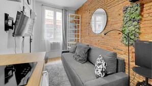 A modern living room with a grey sofa, a flat-screen TV mounted on the wall, a wooden dining table, and a brick wall with a green plant decoration. - Getaway For 2, Beautiful Studio Near The Garonne (Toulouse)