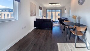 Een moderne woonkamer met een donkere bank, een eettafel met stoelen en een groot raam met jaloezieën.