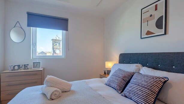 Een keurig opgemaakt slaapkamer met een bed, een nachtkastje met een lamp, een raam met uitzicht op een klokkentoren, en een spiegel aan de muur.