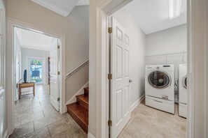 A laundry room with a front-loading washer and dryer, a wooden staircase, and a hallway leading to a room with a television and a view of the outdoors. - Large Laguna Beach Home Across from Beach w/Ground Floor Suite, FREE Perks! (Panama City Beach)
