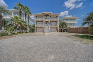 A multi-story beachfront property with a central staircase leading to the entrance, surrounded by palm trees and a wooden fence. - Large Laguna Beach Home Across from Beach w/Ground Floor Suite, FREE Perks! (Panama City Beach)