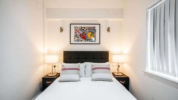 A neatly made bed with white linens and striped pillows, flanked by two bedside tables with lamps, in a room with a large abstract painting on the wall and a window with blinds. - The Anfield Hotel (Liverpool)
