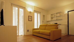 A modern living room with a yellow sofa, wooden flooring, and a kitchen area with white cabinets. - Italianway Easy - Caterina da Forlì 6 (Milan)