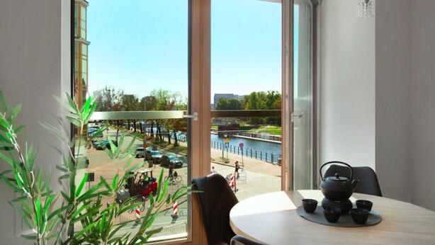 A room with a view of a riverside promenade, a table with a teapot and cups, and a potted plant. - Elite Apartments Waterlane SPA (Gdansk)