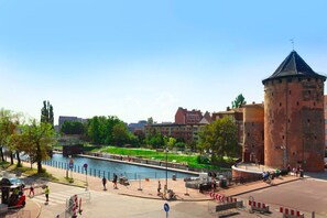 A historic tower with a conical roof, a river in the foreground, and modern buildings in the background.