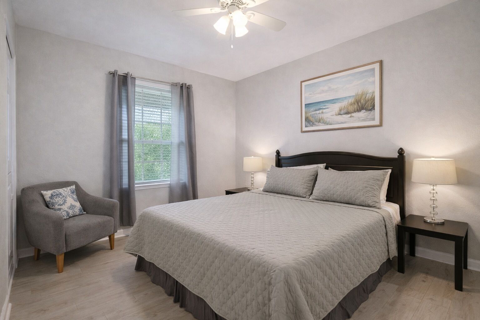 A bedroom with a bed, a chair, a nightstand, a lamp, a window with curtains, a ceiling fan, and a painting on the wall.