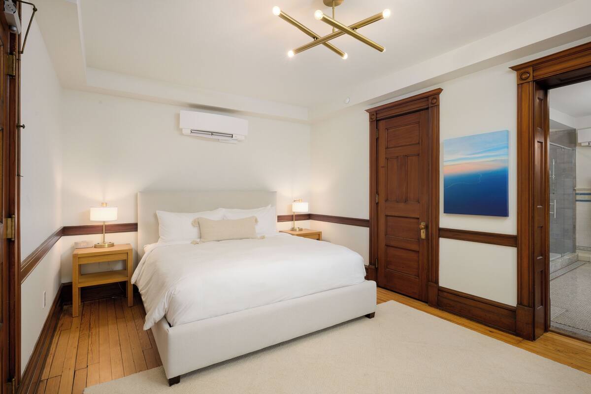 Un dormitorio con una cama grande, mesitas de noche de madera, una lámpara de techo moderna, una puerta de madera y un aire acondicionado instalado en la pared.