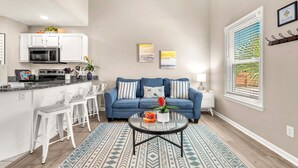 Ein modernes Wohnzimmer mit einem blauen Sofa, weißen Barhocker, einem Glastisch und einer Küchenzeile mit weißen Schränken und einer Mikrowelle.