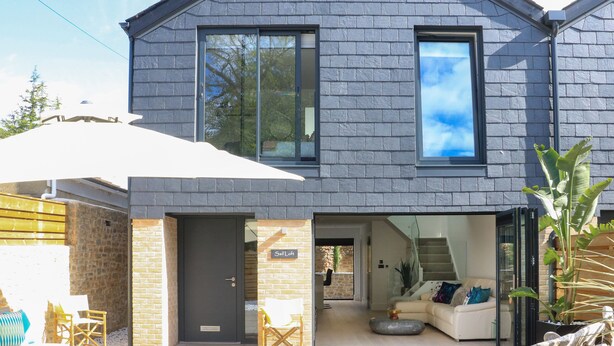 A modern two-story house with a brick exterior, large windows, and a visible interior with a sofa and a staircase. - Sail Loft (Dawlish)