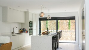 A modern kitchen with white cabinetry, a marble island, and a dining area with bar stools. - Sail Loft (Dawlish)