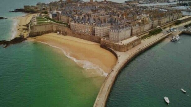 Vista aérea de una ciudad costera con una imponente estructura que parece un castillo, una playa de arena y un mar tranquilo.