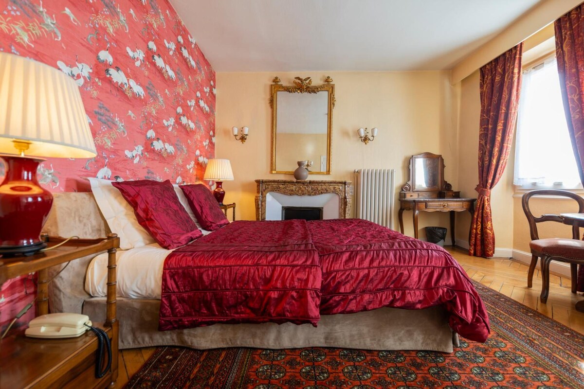 A hotel room with a large bed, a desk, a chair, a radiator, and a window with curtains.