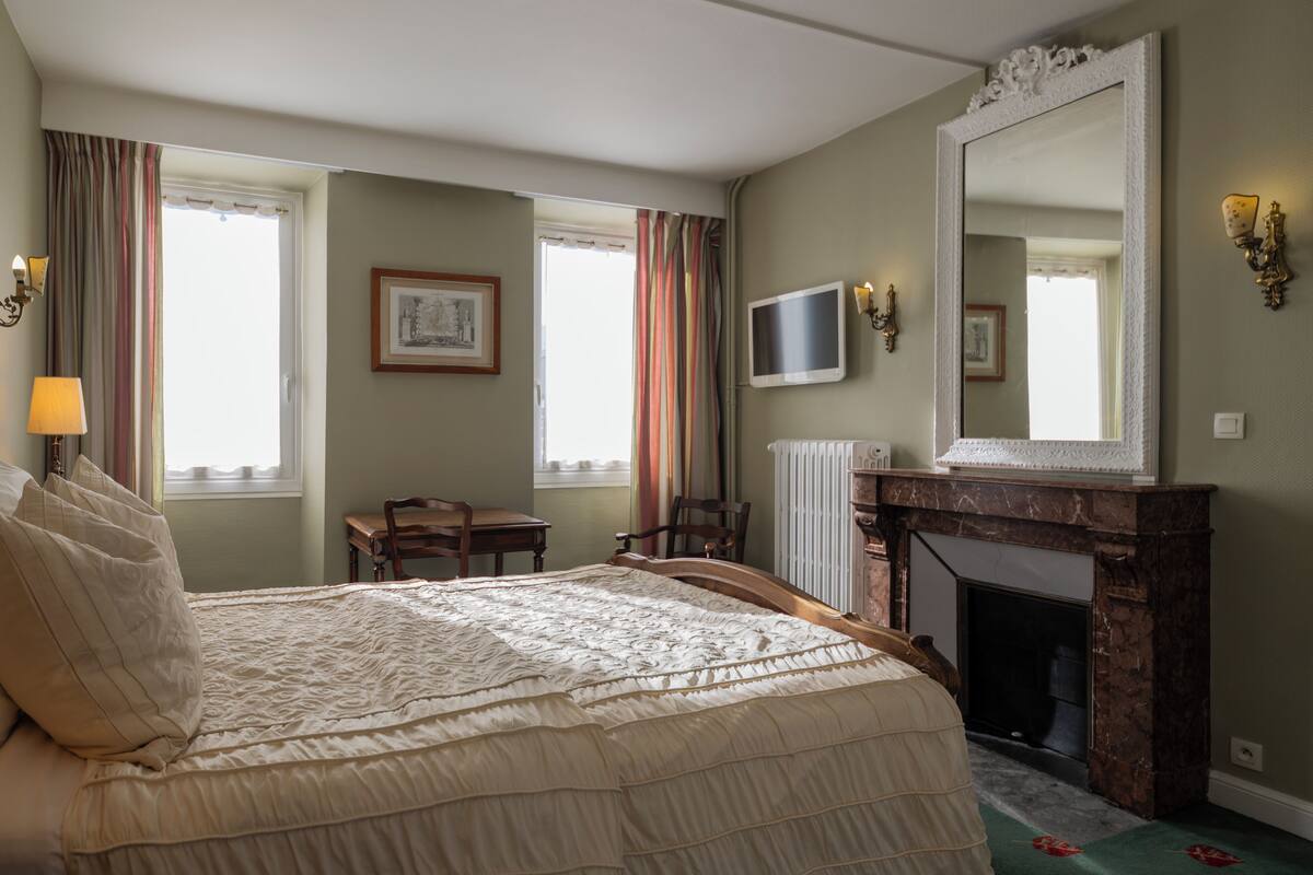 A bedroom with a bed, a chair, a desk, a fireplace, and a television.