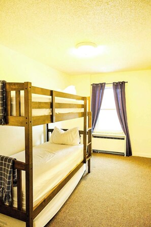 A single bed with a wooden frame, a window with curtains, and a ceiling light.