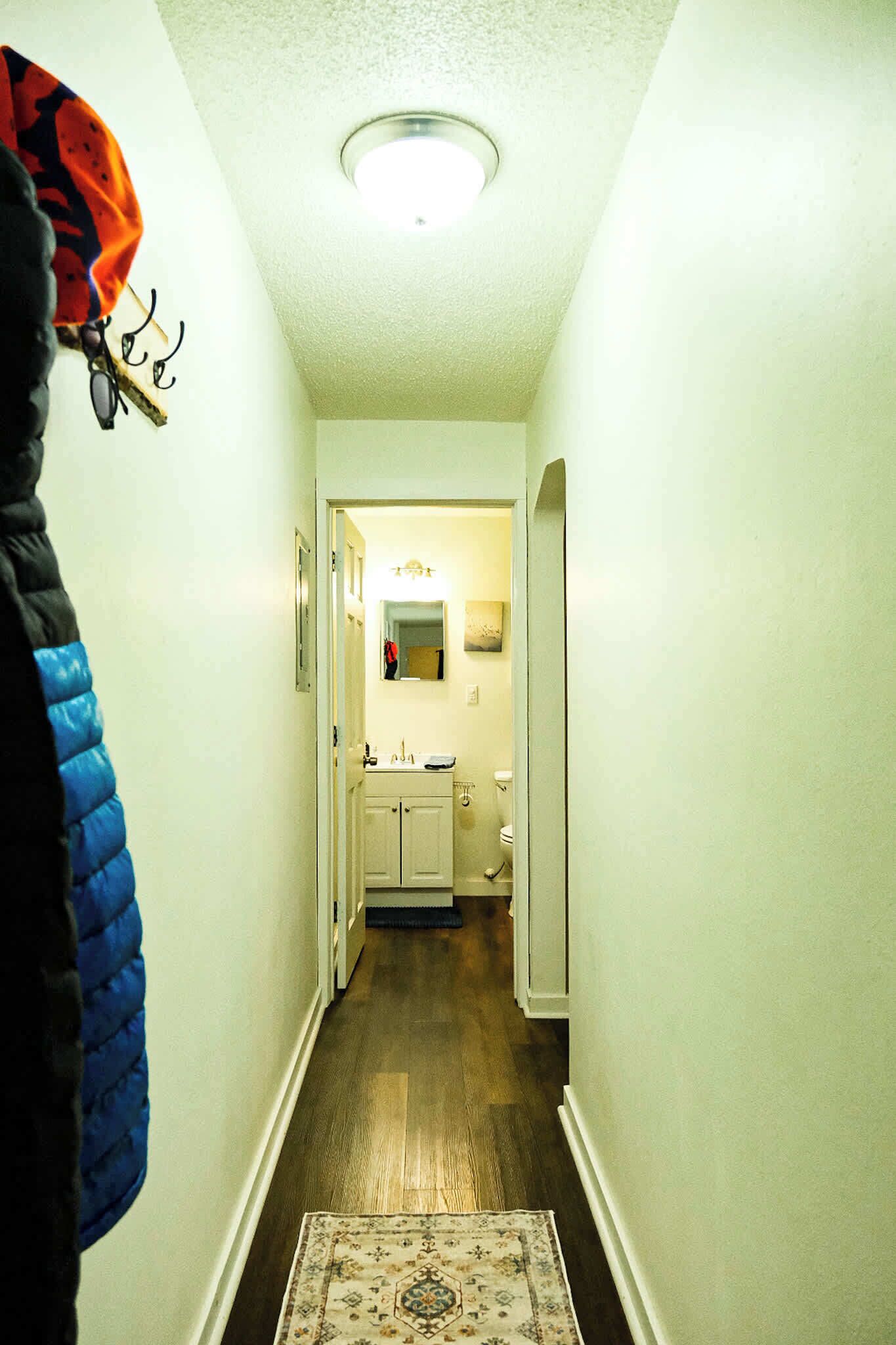 Un couloir étroit, doté d’un plancher en bois, mène à une salle de bain équipée d’un lavabo et de toilettes.