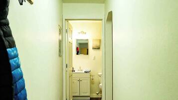 Un couloir étroit, doté d’un plancher en bois, mène à une salle de bain équipée d’un lavabo et de toilettes.