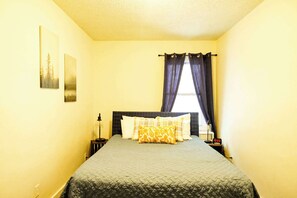 A neatly made bed with a quilted bedspread and multiple pillows, a bedside table with a lamp, and a wall-mounted picture.