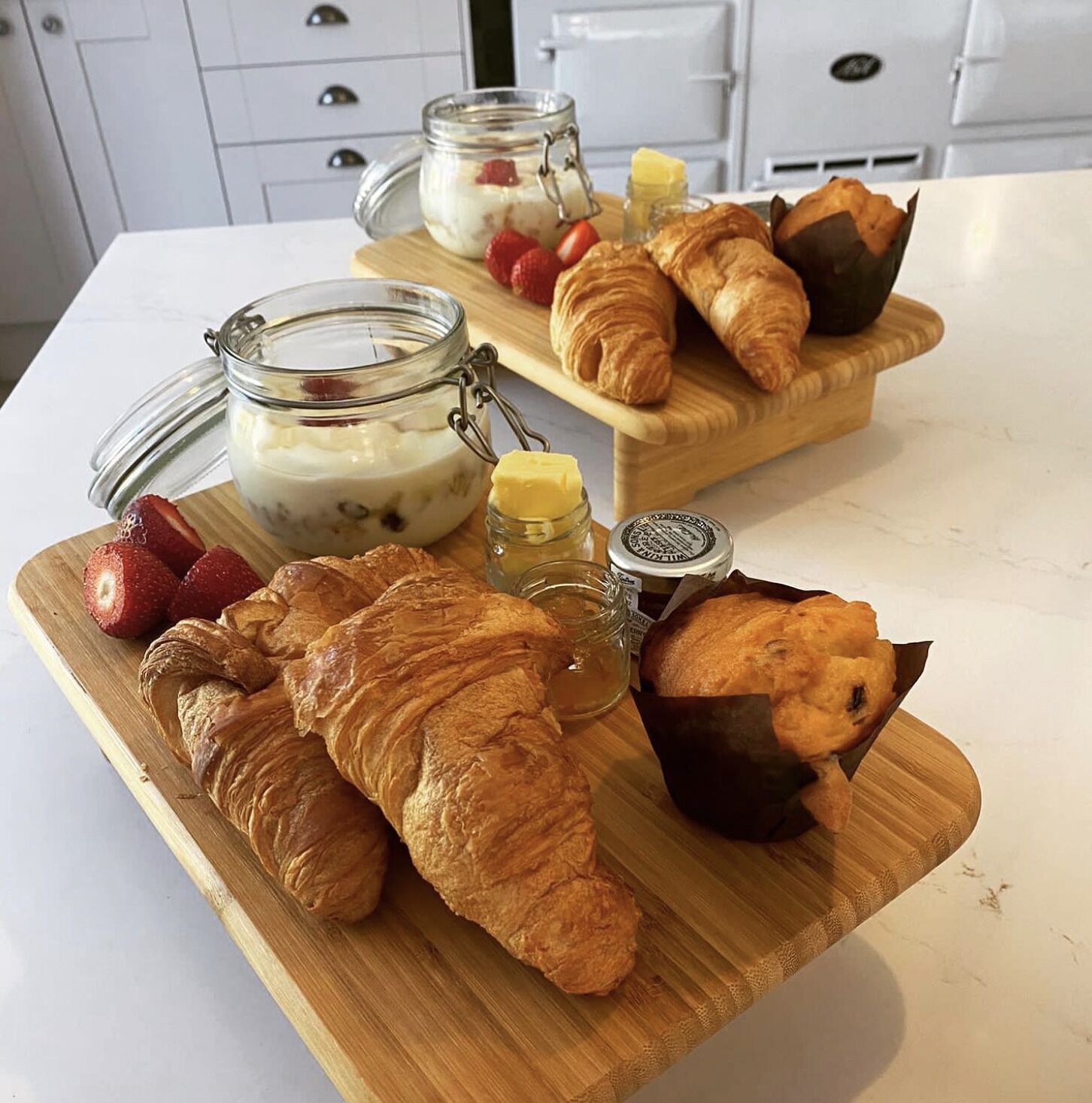 A breakfast setting with croissants, strawberries, and a jar of yogurt parfait on a wooden board.