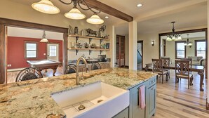 A spacious kitchen with granite countertops, a white sink, and wooden cabinets. There are pendant lights above the sink and a dining area with wooden chairs and a table.