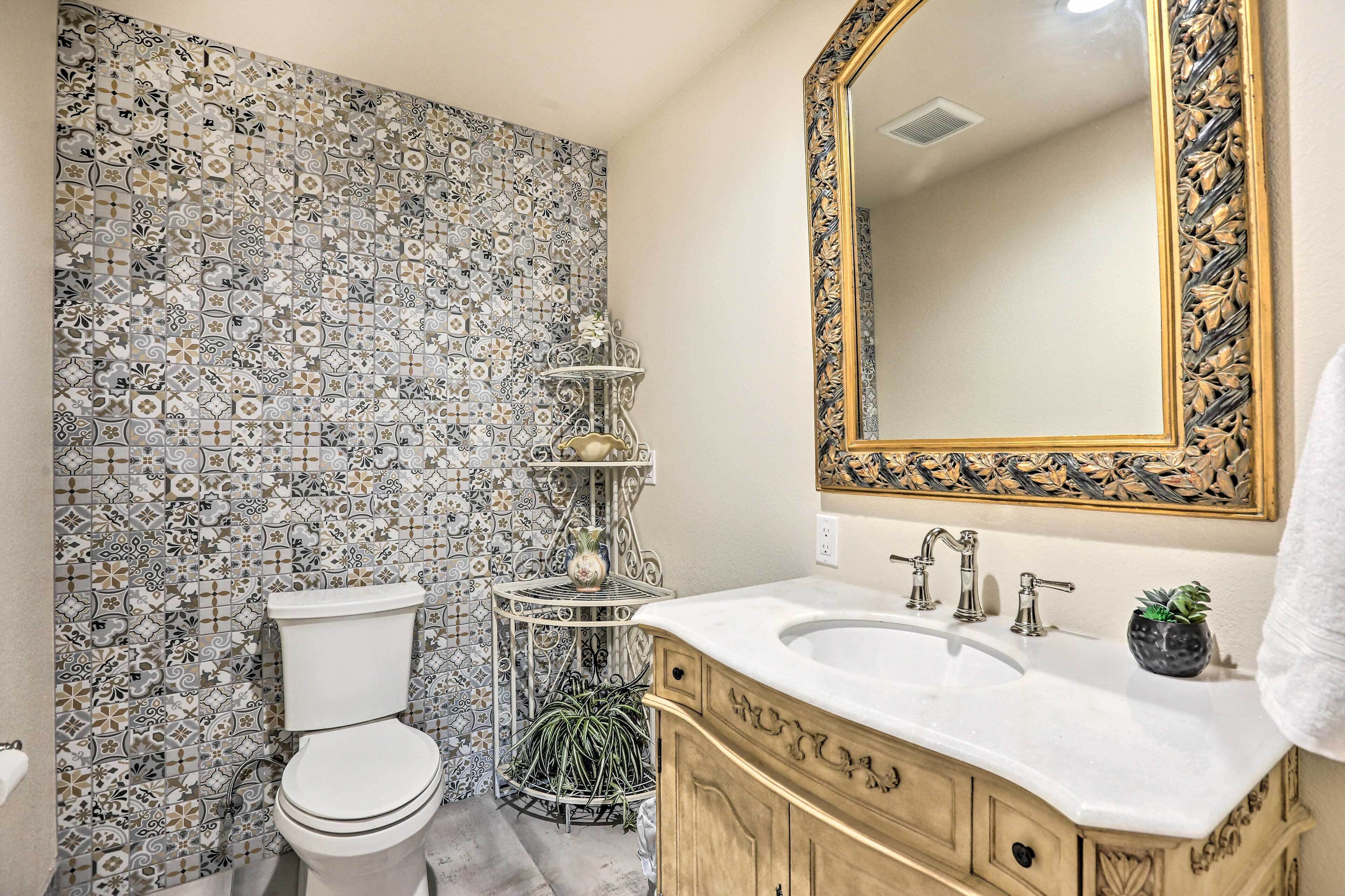 A bathroom with a mosaic tile wall, a white toilet, a vanity with a gold frame, and a large mirror.