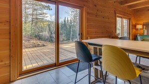 A wooden cabin with a large sliding glass door leading to a deck. Inside, there is a dining table with chairs and a lamp. - L'Anse 750 sur la rivière Sacacomie (Saint-Alexis-des-Monts)