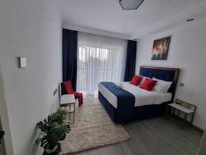 Una habitación de hotel moderna con una cama grande, un escritorio pequeño, una silla y un ventanal con cortinas.