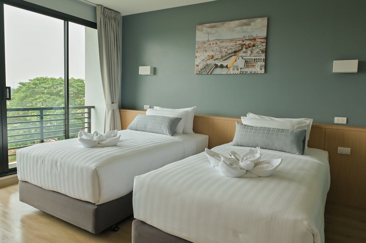Habitación de hotel con dos camas, un cuadro de paisaje urbano en la pared y un balcón con vistas a árboles.