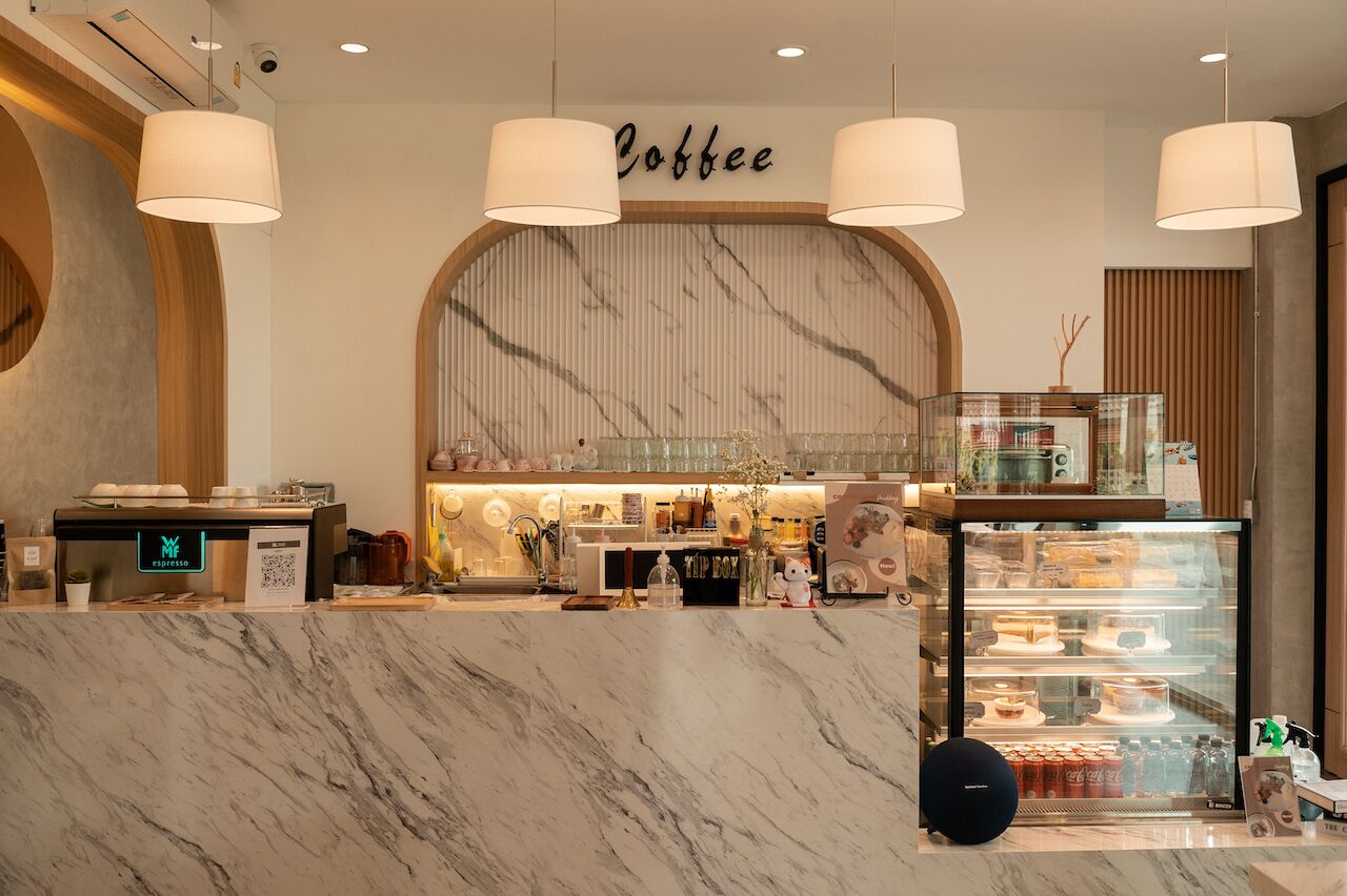 Interior de una cafetería con mostradores de mármol, un letrero de café y una vitrina con pasteles.