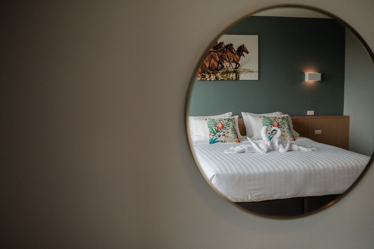 A hotel room with a round mirror reflecting a bed with white bedding and floral pillows, a dark blue wall, and a painting of horses.