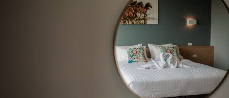 A hotel room with a round mirror reflecting a bed with white bedding and floral pillows, a dark blue wall, and a painting of horses.