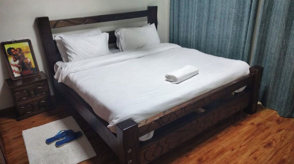 A neatly made bed with white linens, a wooden headboard, and a pair of slippers on a rug.